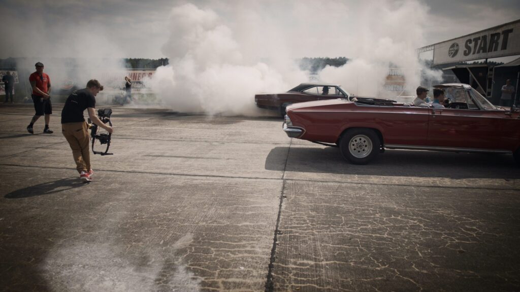 A camera operator films a car performing a burnout on a track, producing thick white smoke. Two classic convertible cars are parked nearby, with people seated inside. The start line is visible in the background under cloudy skies.