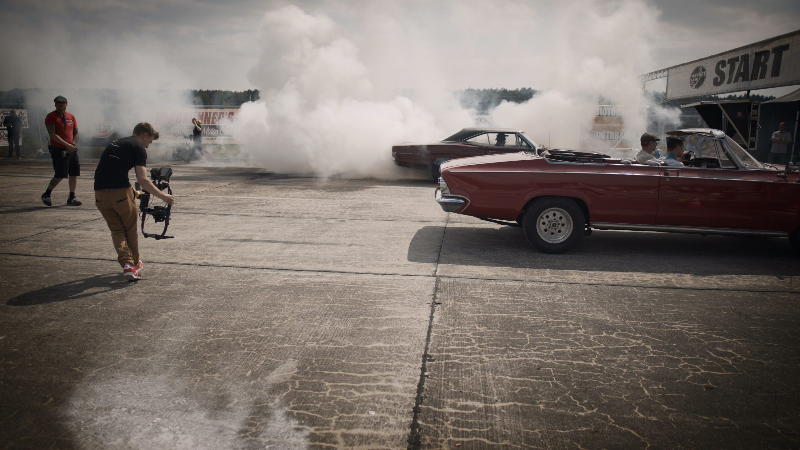 A camera operator films a car performing a burnout on a track, producing thick white smoke. Two classic convertible cars are parked nearby, with people seated inside. The start line is visible in the background under cloudy skies.