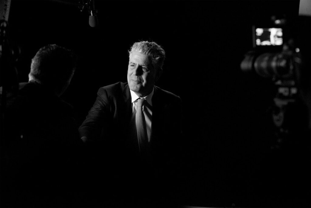 Black and white image of a man in a suit sitting in a dimly lit setting, facing another person. He appears thoughtful, with studio equipment visible around them.