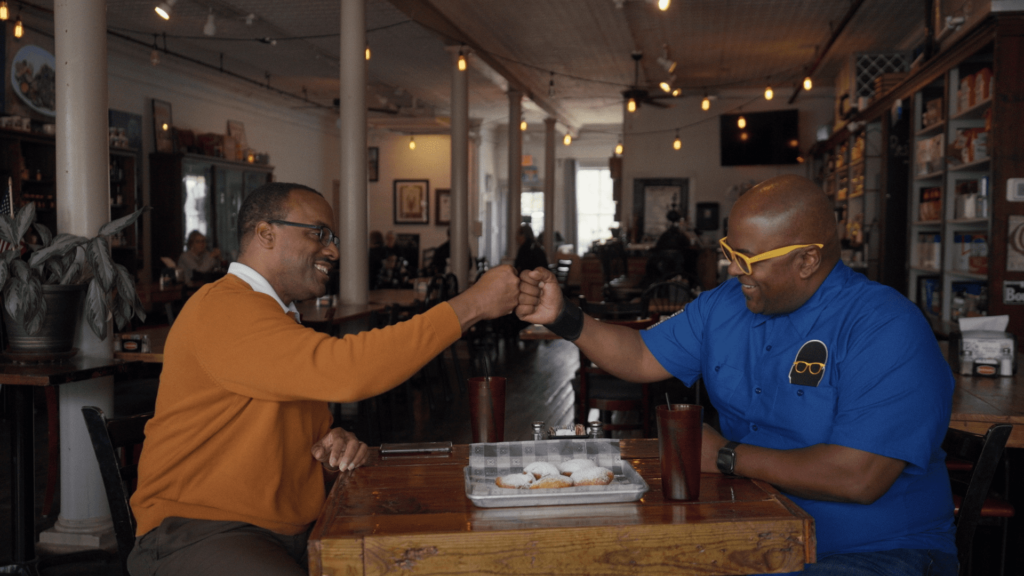 Two people sitting across a wooden table in a cozy cafe, smiling and bumping fists. One wears an orange sweater, the other a blue shirt with yellow glasses. Plates and drinks are on the table, and the background shows shelves and wall decorations.