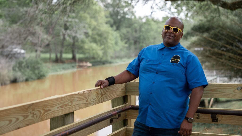 Smiling man in a bright blue shirt and yellow sunglasses leans casually against a wooden railing. Behind him, a calm, brownish river flows through a lush, green, wooded area.