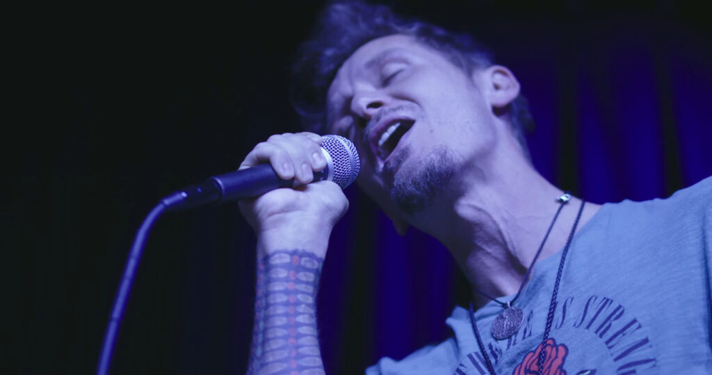 A person passionately sings into a microphone on stage, eyes closed, with an expressive face. They have a tattooed arm, wear a necklace, and the lighting casts a cool blue tone over the scene.