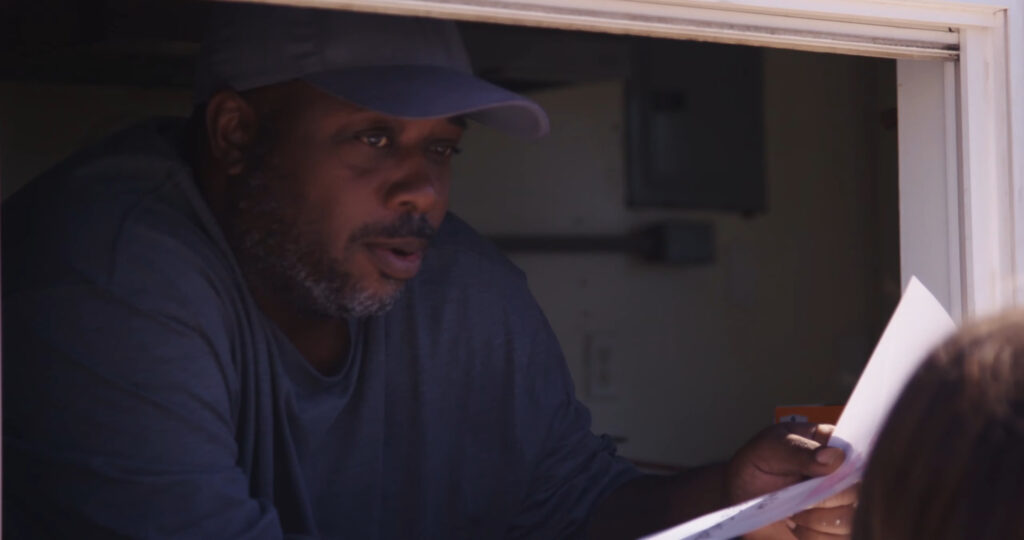 A man in a cap leans out of a window, holding papers. He appears to be in conversation with someone outside. The interior behind him is dimly lit, suggesting he might be in a small booth or vehicle.
