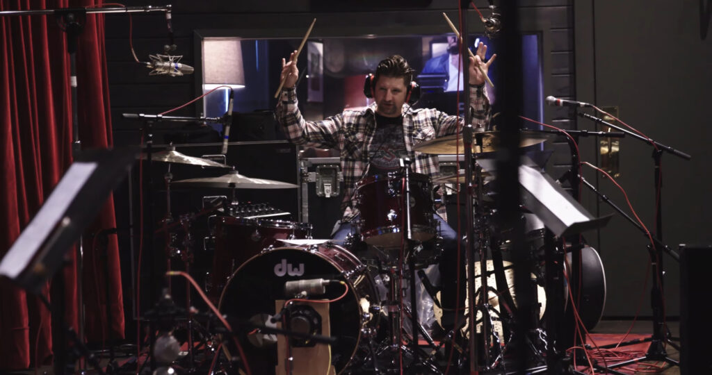 A drummer in a plaid shirt energetically playing a drum set in a recording studio. Various microphones and cables surround the drums. The drummer is raising the drumsticks with both hands.