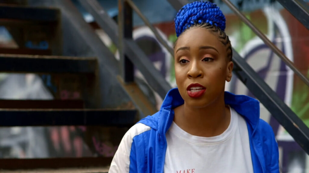 Person with blue braided hair in a bun, wearing a blue jacket over a white t-shirt, is speaking. They are outside with blurred stairs and graffiti in the background.