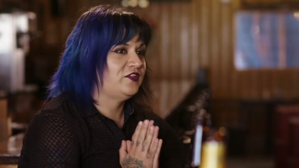A person with blue hair and a black outfit sits at a table with a wooden background. They have their hands together, showing a tattoo on one hand, and appear to be engaged in conversation. Two bottles are visible in the foreground.