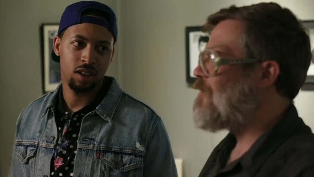 Two men are indoors. One wears a blue cap, a denim jacket, and a patterned shirt. The other has glasses, a beard, and a dark shirt. They seem to be having a conversation. A framed picture is on the wall behind them.