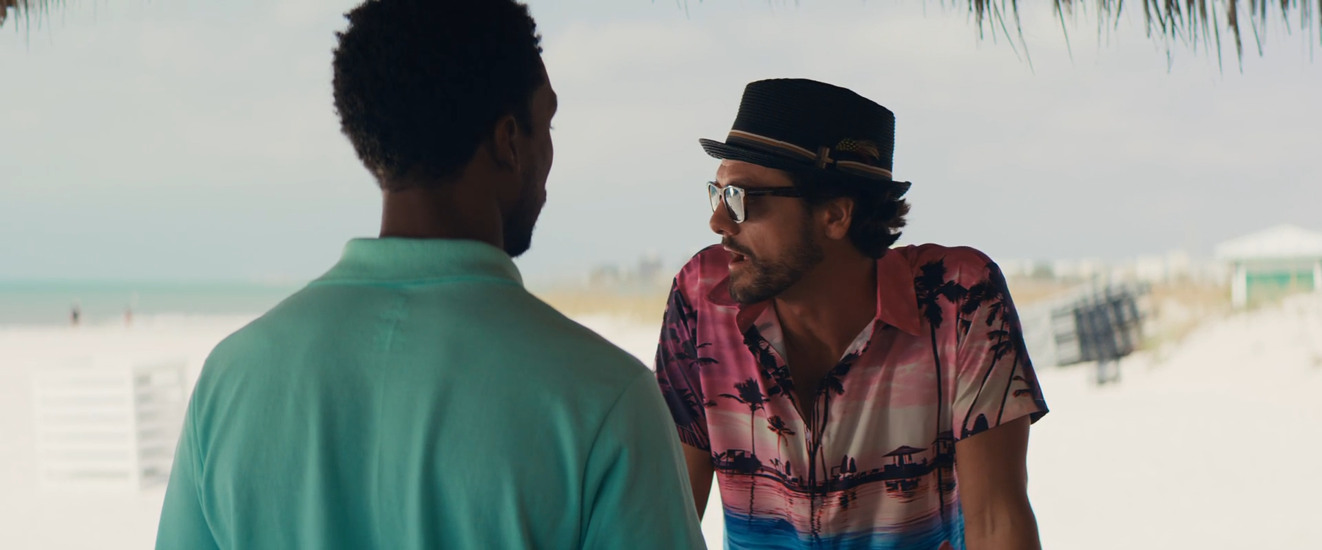 Two men are talking at a beach. One man wears a turquoise shirt and faces away from the camera. The other wears a colorful shirt, glasses, and a hat, and is leaning on a surface. The beach and ocean are blurred in the background.