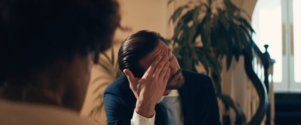 A man in a suit sitting indoors covers his face with one hand, looking stressed. A blurred figure is in the foreground, and a large plant and staircase are visible in the background.