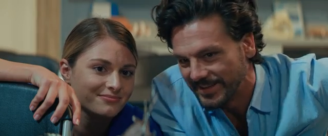 A man and a woman are sitting closely together, both smiling softly. The woman has her head resting on her hand, while the man leans in slightly. They are indoors, with blurred shelves in the background.