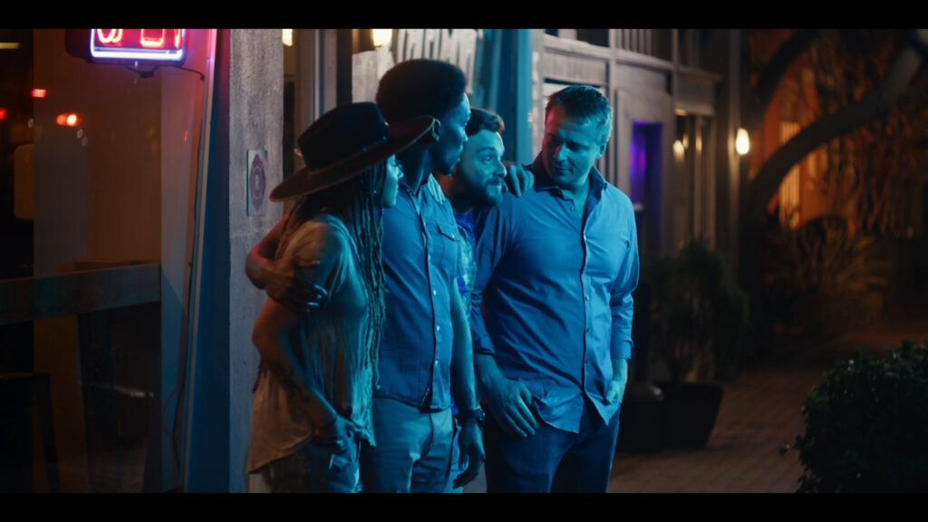 A group of four people stand together on a dimly lit street at night, engaged in conversation. They are outside a building with colorful neon lights, wearing casual clothes. One person wears a hat, and they all appear relaxed and friendly.