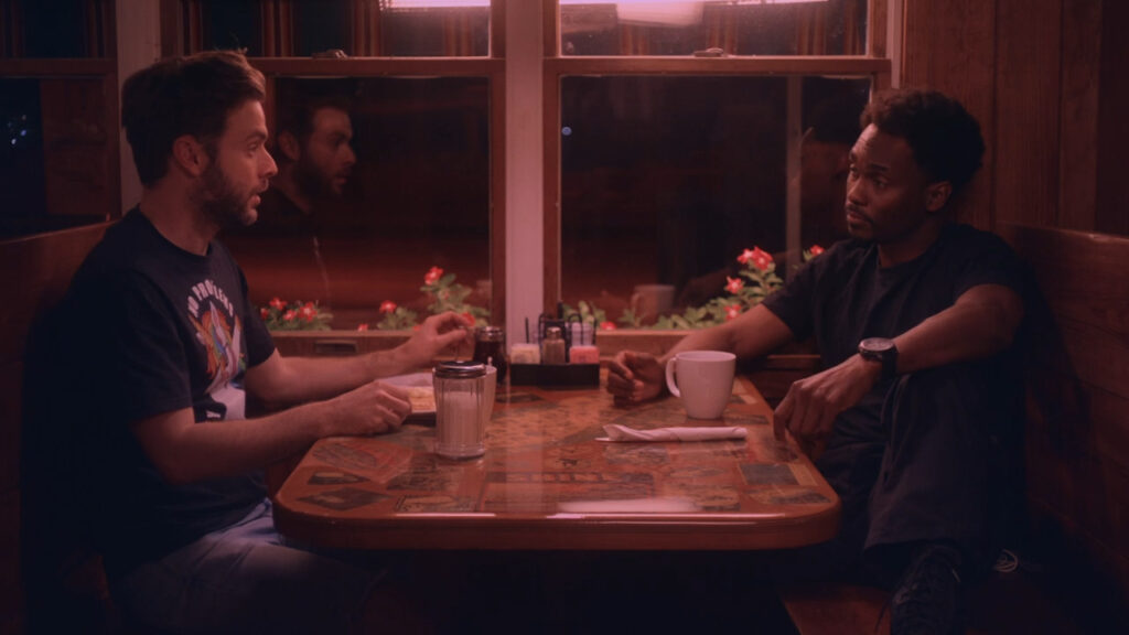 Two men are sitting across from each other in a dimly lit diner booth. One is gesturing while talking, and the other is listening with a cup in front of him. The table has salt and pepper shakers and a vase with red flowers by the window.