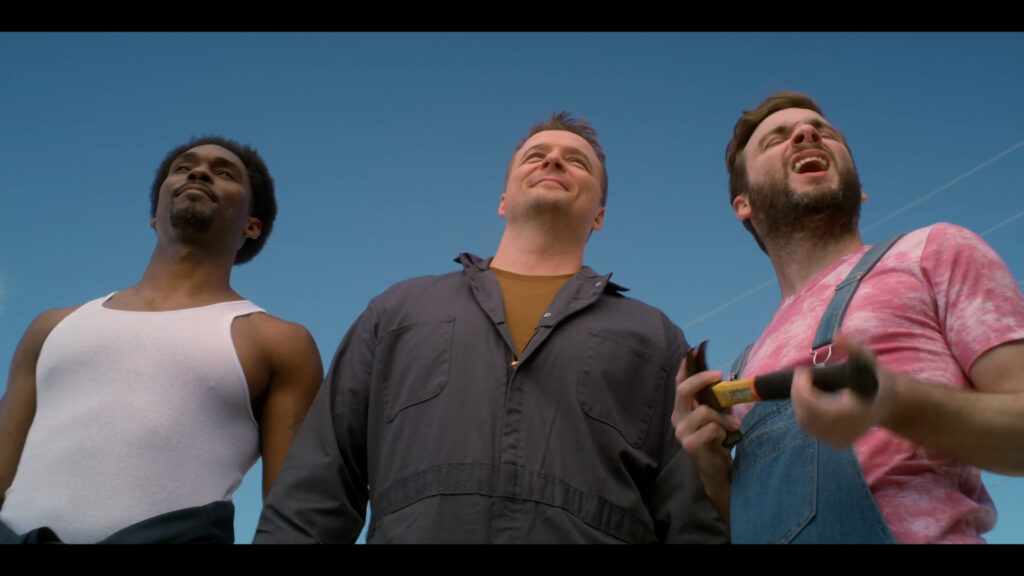 Three men stand outside against a clear blue sky, smiling and looking upwards. The man on the left wears a white tank top, the middle man wears a dark jumpsuit, and the man on the right wears denim overalls and holds a hammer.