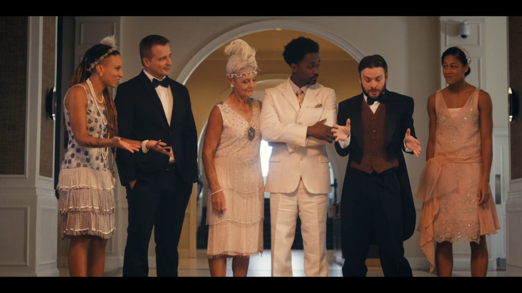 A group of six people dressed in formal vintage attire stand in a well-lit room. The two men wear suits; one in a black suit gesturing animatedly. Two women wear flapper dresses. They appear engaged in conversation, with intricate decor in the background.
