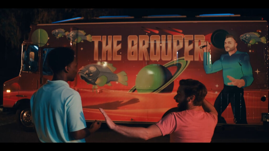 Two people stand beside a food truck with a vibrant space-themed design. The truck displays "THE GROUPER" in bold letters, featuring planets, a fish in a spacesuit, and an astronaut. The scene is set at night with a warm glow.