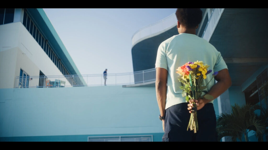 A person stands outdoors, holding a bouquet of colorful flowers behind their back. They face a modern building with curved architecture. Another person is seen at a distance on an upper level walkway. The sky is clear and blue.