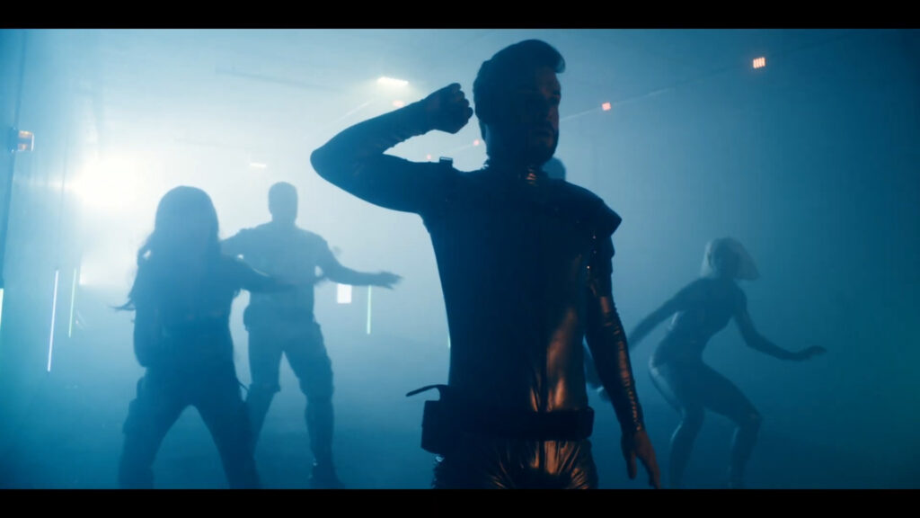 A group of people dancing energetically in a dimly lit, foggy room. Silhouettes are visible due to backlighting, with the central figure in a leather outfit striking a pose. Others are in dynamic positions, adding to the lively atmosphere.