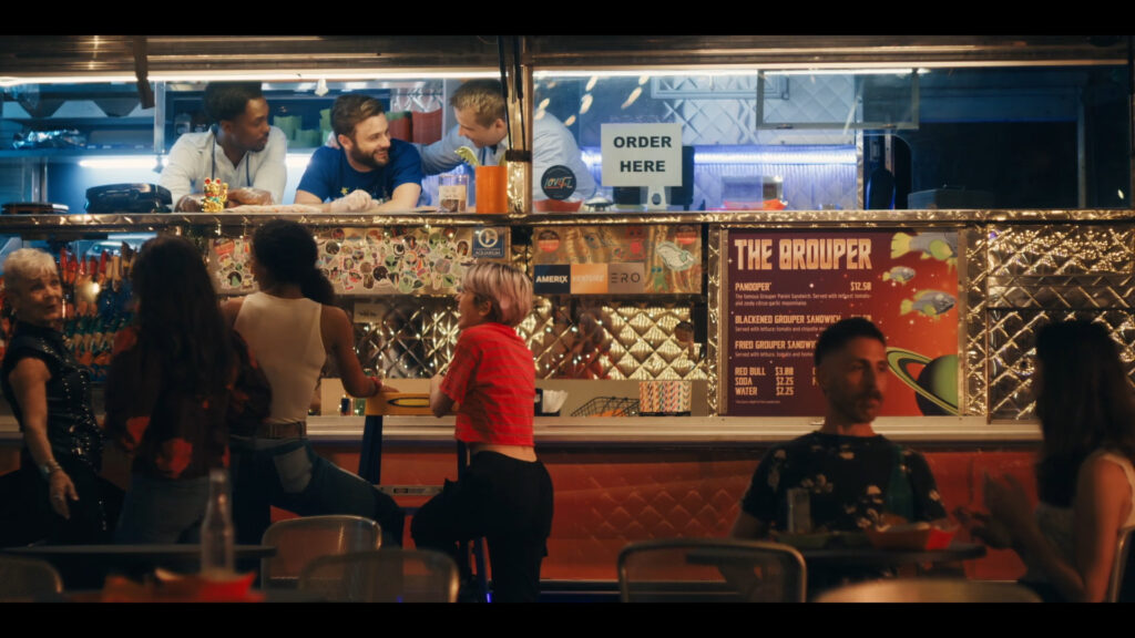 People gather at a food truck at night. Two men inside serve customers while chatting. A group stands outside waiting, including a woman in a red top. The truck has a colorful menu sign, and the area is lively and bustling.