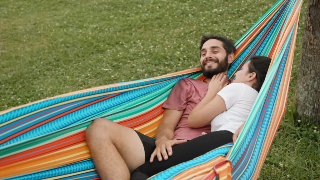A man and a woman relax together in a colorful hammock. The man is wearing a pink shirt and shorts, while the woman, in a white shirt, rests her head on his chest. They are outdoors on a grassy area, looking content and happy.