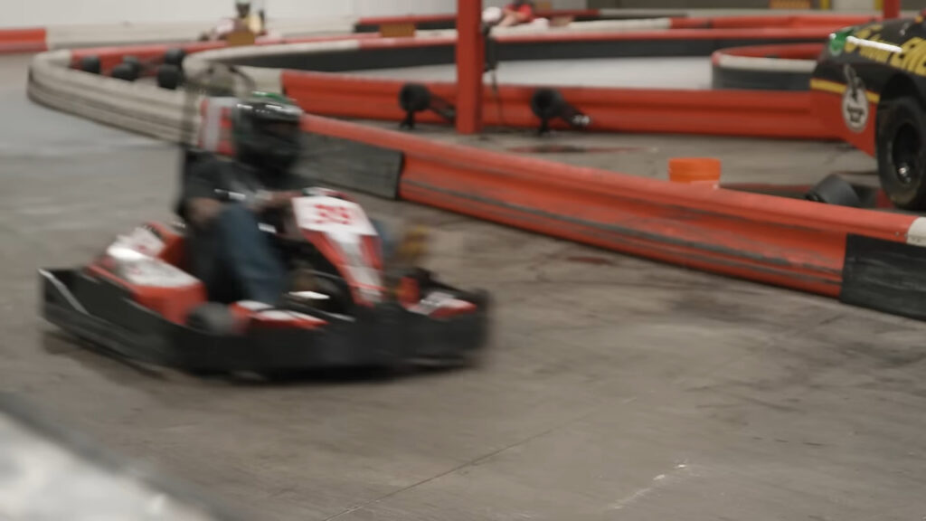 Person driving a go-kart on an indoor track with red and black barriers. The driver is wearing a helmet and the kart has the number 59.