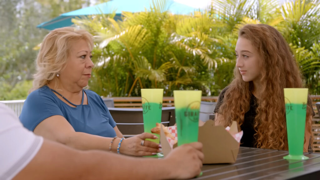 Two women sit at an outdoor table, engaged in conversation. They have long hair and wear casual clothing. Bright green drink cups and an empty food container are on the table. Lush greenery decorates the background.