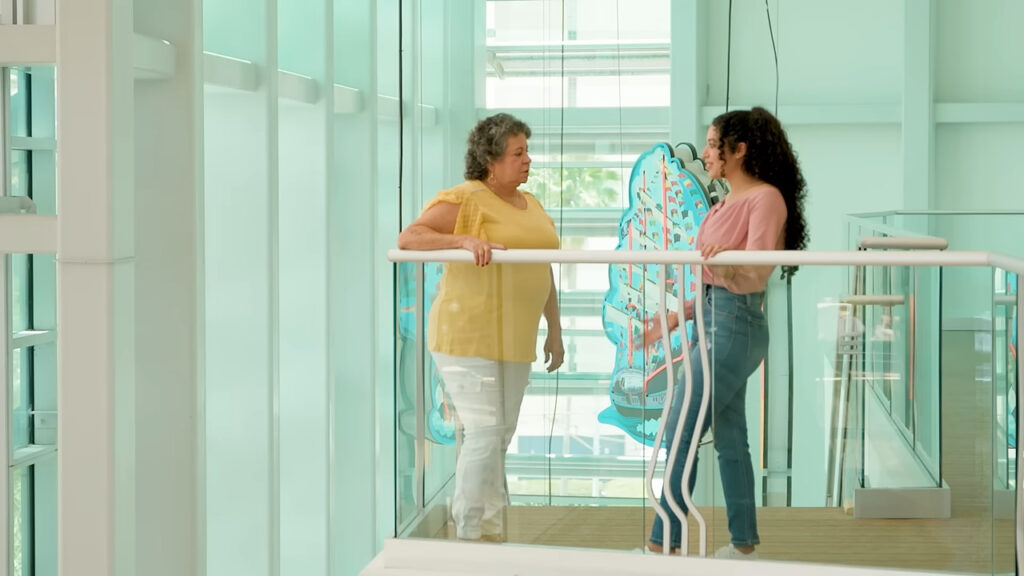 Two women stand and converse on a glass walkway indoors. One wears a yellow top and white pants, while the other wears a pink top and jeans. They are surrounded by large blue butterfly decorations and bright natural light filters through the windows.
