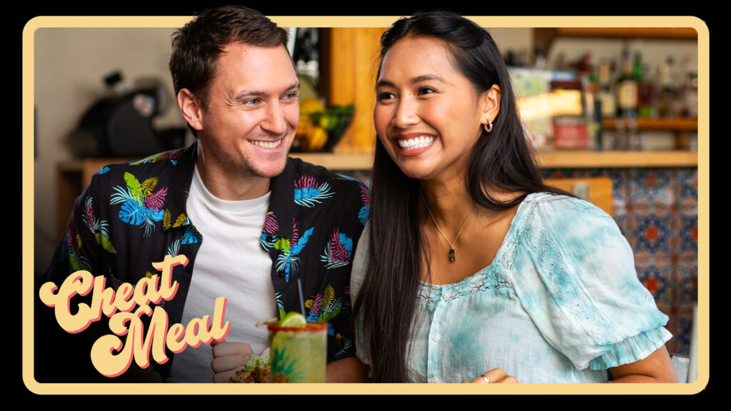 A man and woman are sitting together at a table, smiling. The man wears a black shirt with colorful butterflies, and the woman wears a light blue top. A drink is on the table. The text "Cheat Meal" is in the lower left corner.