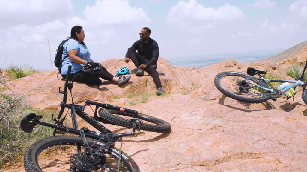Two people resting on a rocky hilltop, with two mountain bikes lying nearby. One is sitting with a helmet beside them, while the other is seated, both engaged in conversation. The sky is partly cloudy.