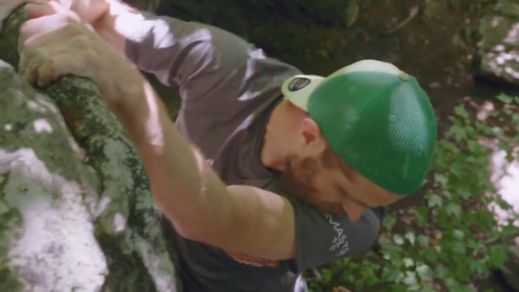 A man wearing a green cap and gray t-shirt is rock climbing outdoors. He is gripping a rock with both hands and focusing intently. Sunlight filters through the surrounding foliage.