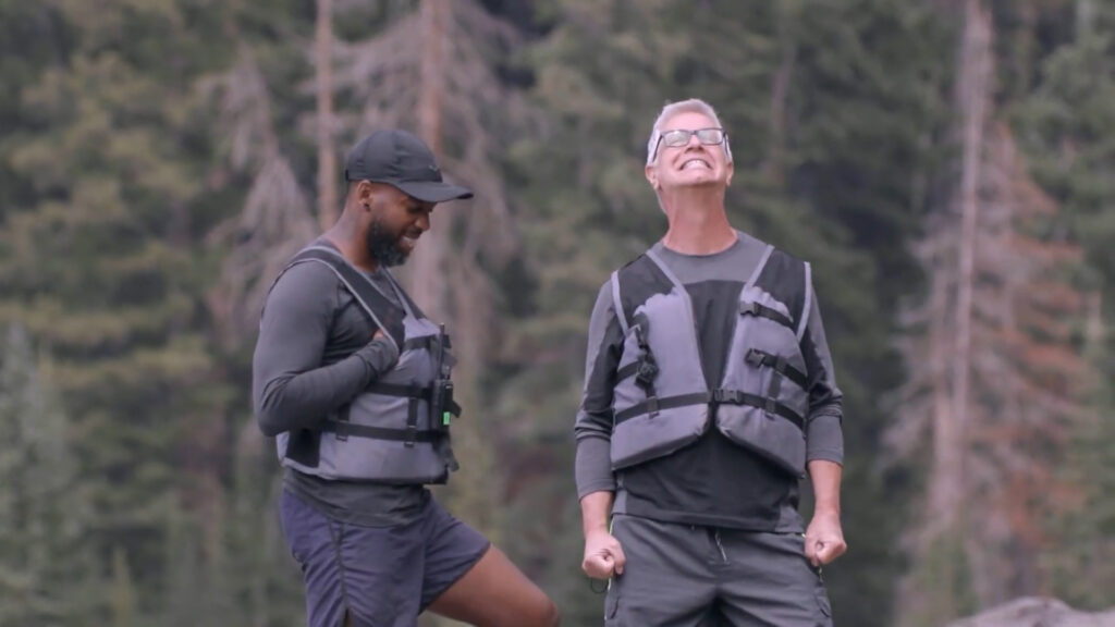 Two people wearing life vests stand outdoors in front of trees. The person on the left is smiling with a binocular harness, while the person on the right is grinning broadly, fists clenched in excitement. Both are dressed in casual outdoor attire.