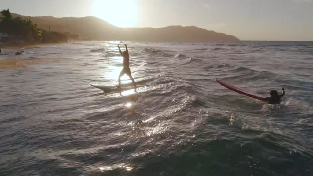 A person surfs on a wave at sunset near a tropical beach, with arms raised in excitement. Another person is in the water nearby, holding onto a red surfboard. The sun sets behind distant hills, casting a warm glow over the scene.