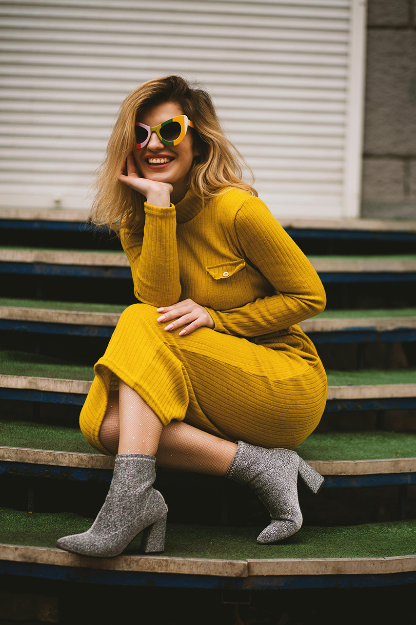 A woman in a yellow dress sits on steps, smiling. She rests her chin on her hand and wears reflective sunglasses and sparkly silver ankle boots. The background includes a closed shutter.