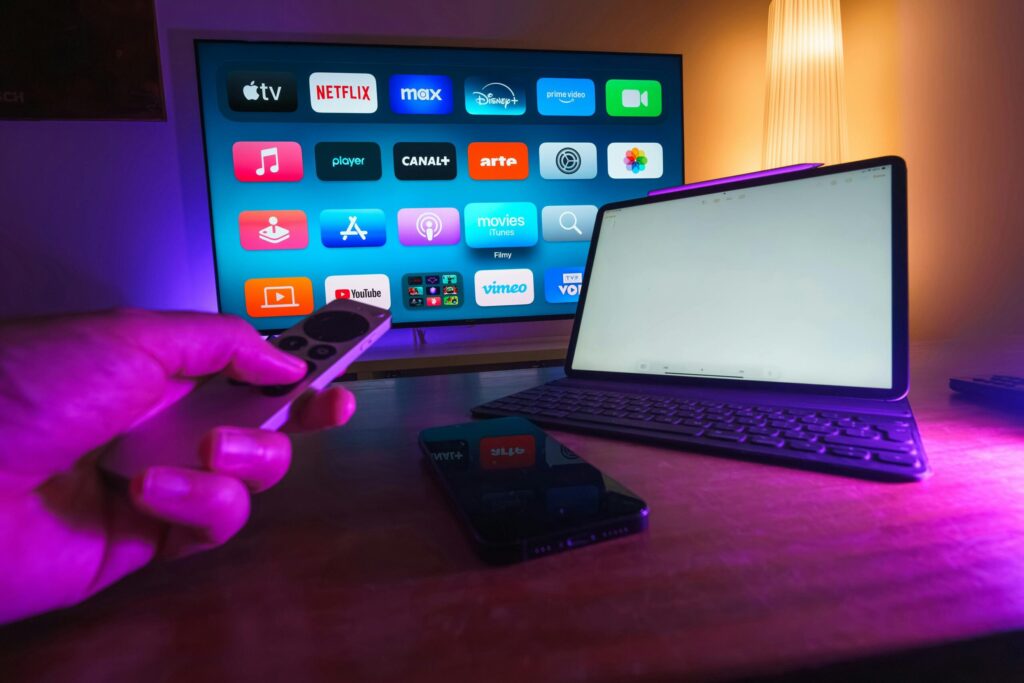 A hand holds a remote control in front of a TV screen displaying various streaming service logos like Netflix and HBO Max. Nearby, a tablet with a keyboard and a smartphone are on a table, all illuminated by purple ambient lighting.