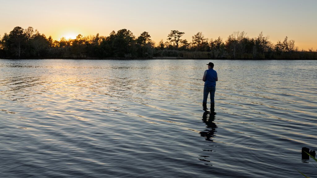 A person standing on a calm lake at sunset, wearing casual clothes. The sun sets behind a treeline in the distance, casting a warm glow over the water. The person appears to be fishing, with trees lining the background.
