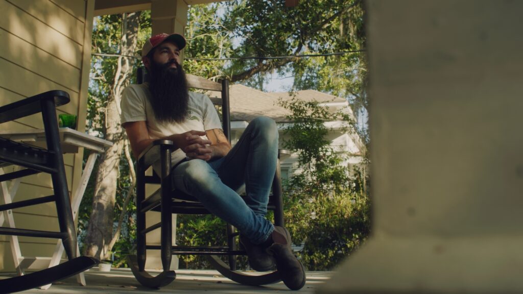 A man with a long beard, wearing a red cap, white t-shirt, and jeans, sits relaxed in a rocking chair on a shaded porch surrounded by trees and greenery.