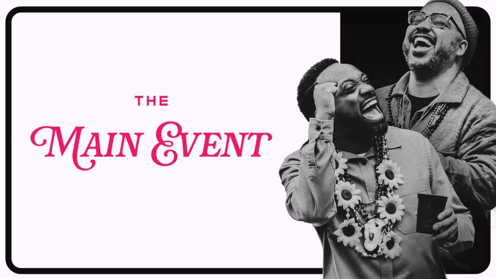 Black and white image with two men laughing and wearing floral necklaces. Text reads "The Main Event" in pink, with "The" in smaller uppercase letters above "Main Event" in bold script.